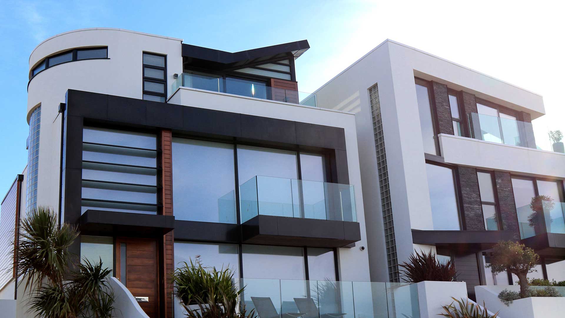 Modern two-story house with large glass windows and geometric design, offering stunning views from the balconies. Surrounded by minimal landscaping, it stands proudly against a bright blue sky. Its impressive scale is sure to capture attention and inspire any marketing showcase.