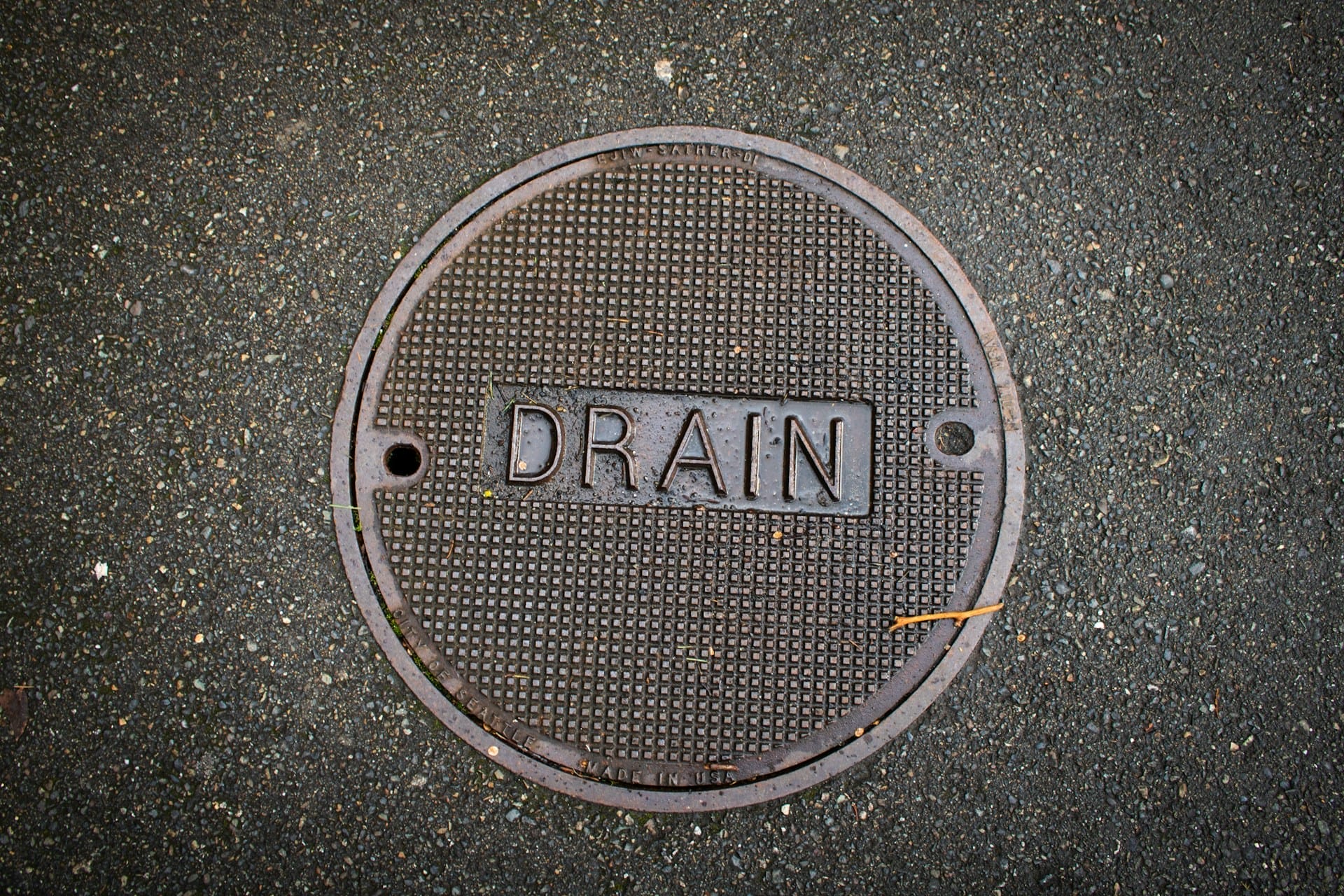 A round metal manhole cover labeled "DRAIN" set in an asphalt surface, symbolizing how poor small business marketing waste can drain your resources without cost-effective digital marketing or smart marketing budget optimization.